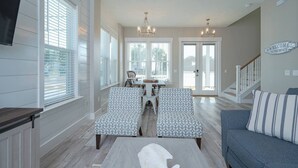A spacious living room with a gray sofa, patterned chairs, a dining table, and a kitchen area with a TV. - Sandy of Panama City Beach - Ocean View, Heated Pool, Steps to Beach, Sleeps 18 (Panama City Beach)