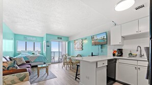 A modern kitchen with white cabinets and a central island, a dining area with wooden chairs, a living room with a sofa and a coffee table, and a balcony with a view.