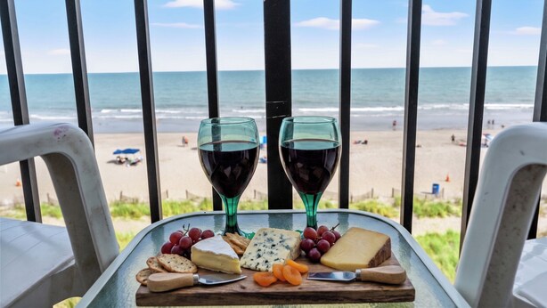A table with cheese, grapes, crackers, and wine glasses on a balcony overlooking a beach.