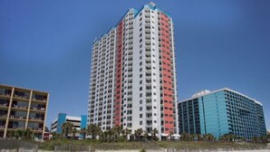 A tall, multi-story hotel with balconies, located near a beach.