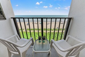 Ein Balkon mit zwei Stühlen und einem Tisch, der auf einen Strand mit Sonnenschirmen und Menschen blickt.