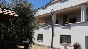 Un edificio blanco con techo de tejas, un balcón con macetas y un árbol grande al frente.