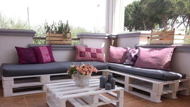 Un rincón exterior acogedor con un banco y una mesa, rodeado de plantas en macetas y cojines decorativos.