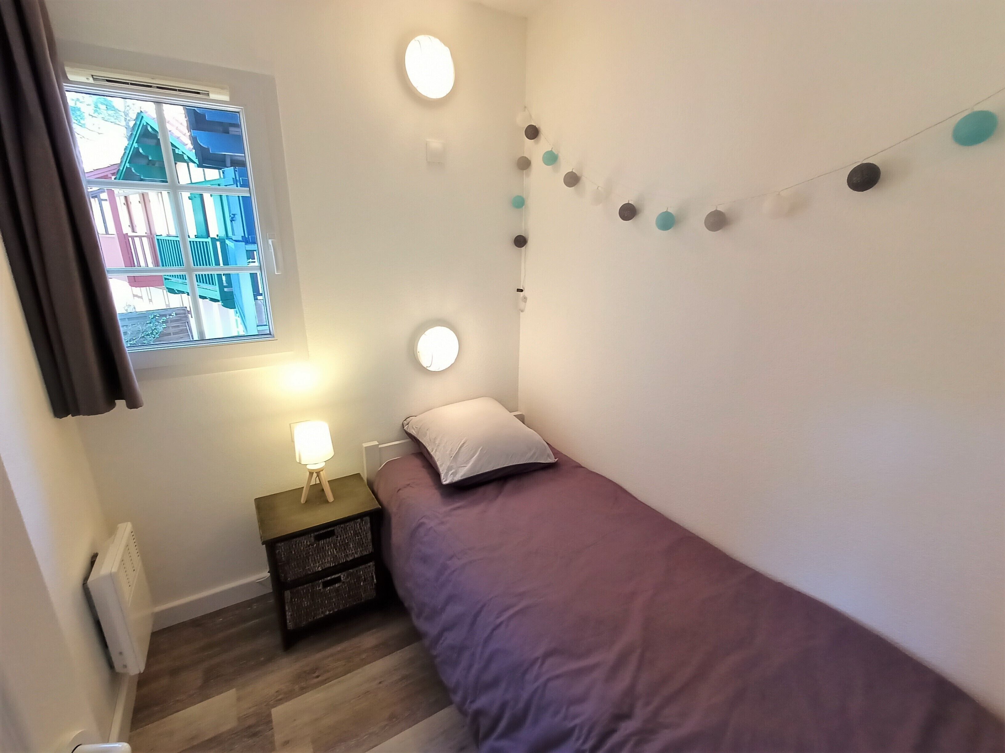 A small, single-bed room with a window, bedside lamp, and wall-mounted light.