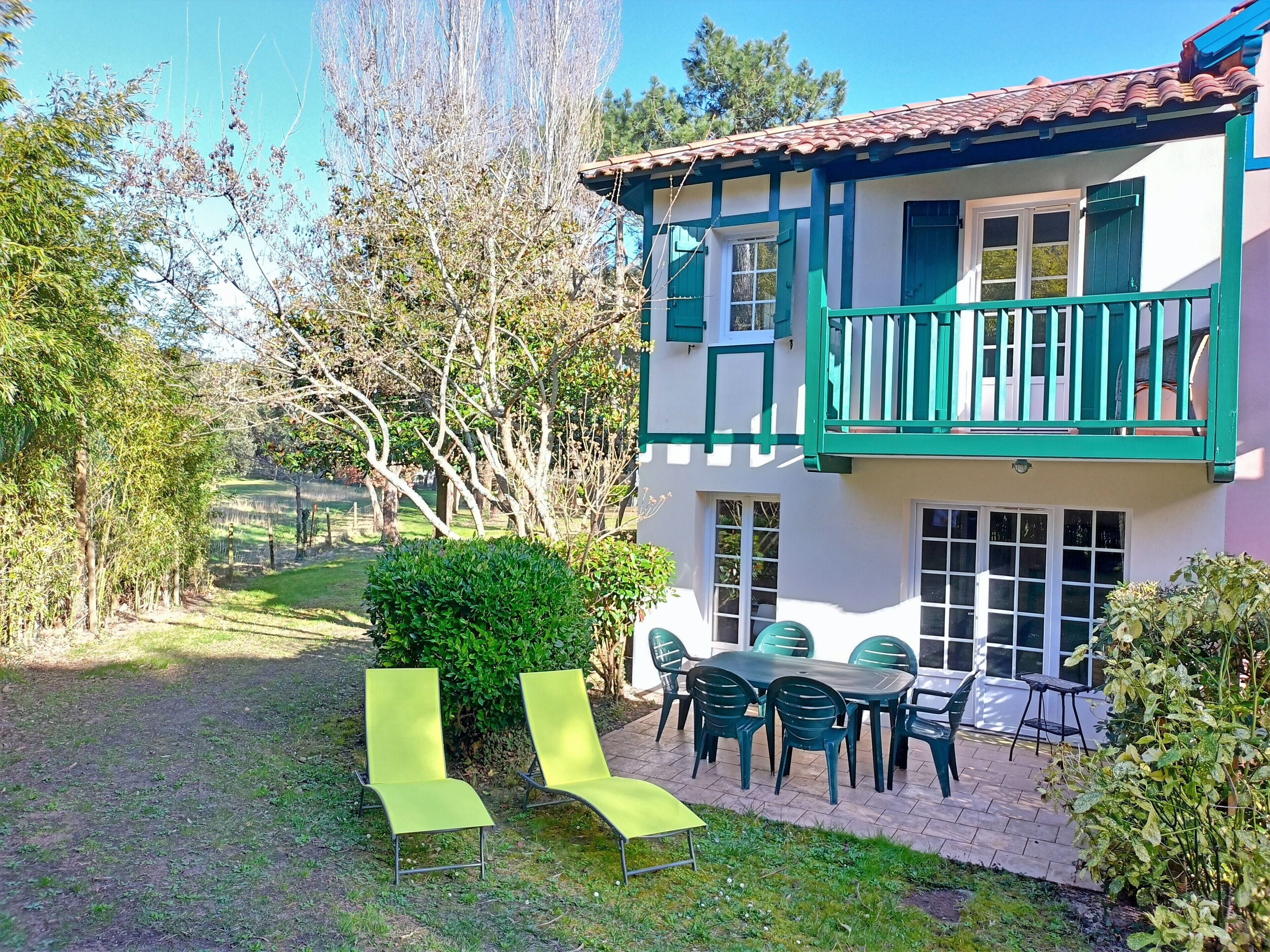 A two-story house with a balcony, a patio with chairs, and a garden.