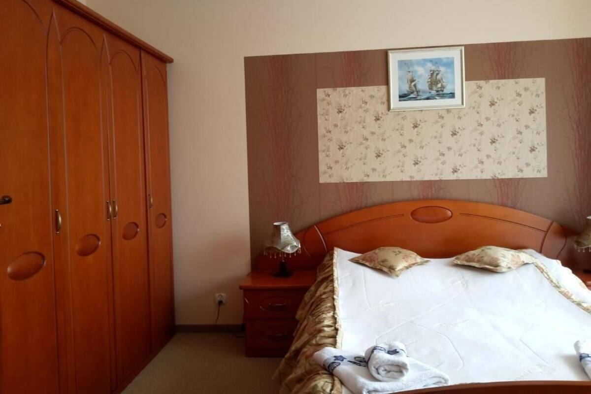 Ein Schlafzimmer mit einem Holzschrank, einem Bett mit Blumenmuster am Kopfteil, einem Nachttisch mit Lampe und einem gerahmten Bild an der Wand.