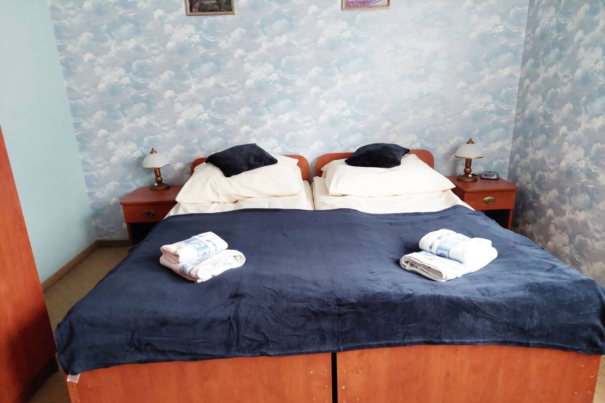 Ein Doppelbett mit dunkelblauer Bettdecke, zwei Kissen und gefalteten Handtüchern. Zwei Nachttische mit Lampen und einer kleinen Uhr stehen bereit. Die Wand ist hellblau und zeigt ein Wolkenmuster.