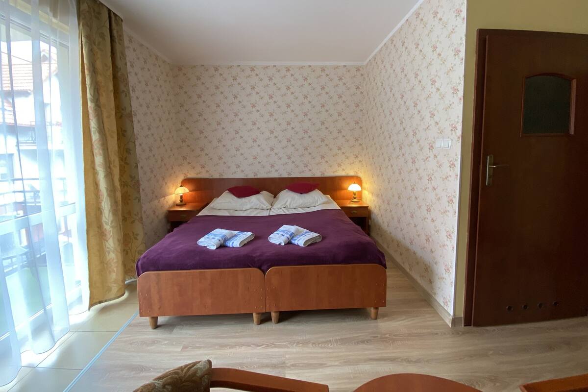 Ein Schlafzimmer mit einem hölzernen Bett, zwei Nachttischlampen, floraler Tapete und einer Holztür.