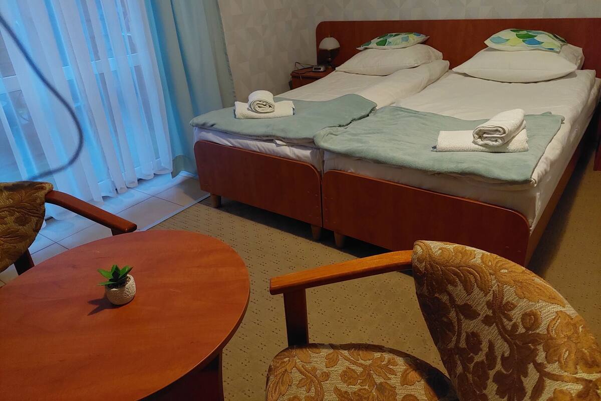 Ein Hotelzimmer mit einem Doppelbett, zwei Kissen, einem Nachttisch mit Lampe sowie einem kleinen Tisch mit Pflanze.