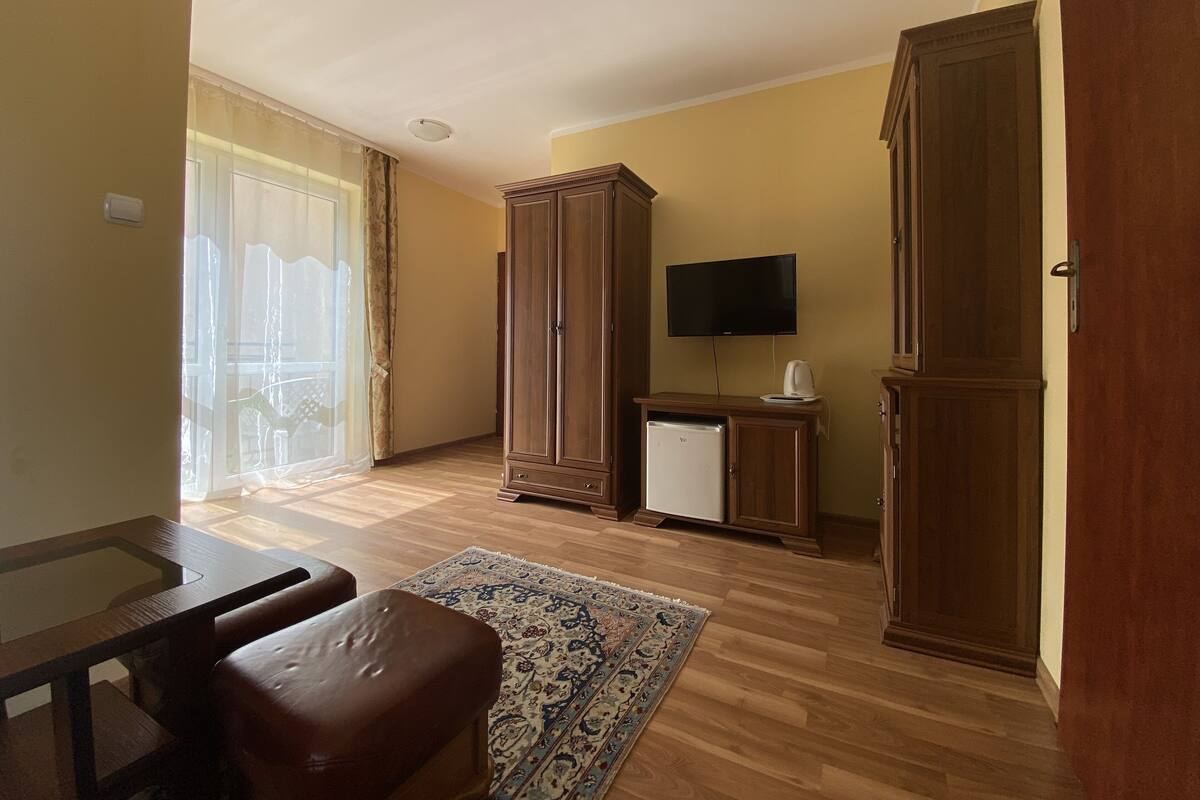 Ein Zimmer mit Holzboden, einem großen Kleiderschrank, einem Fernseher, einem kleinen Kühlschrank, einem Tisch mit Stuhl und einem gemusterten Teppich.