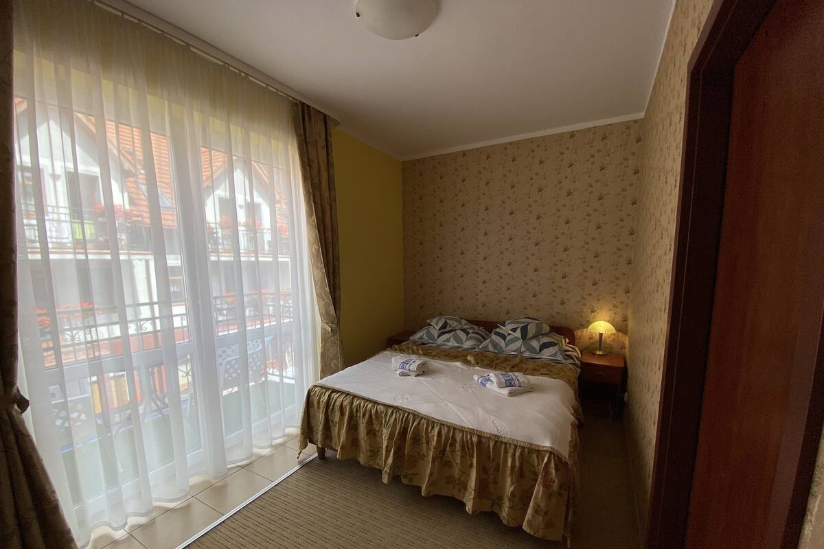 Ein Hotelzimmer mit einem Bett, einem Nachttisch, einer Lampe, einem Fenster mit durchsichtigen Vorhängen und Blick auf einen Balkon.