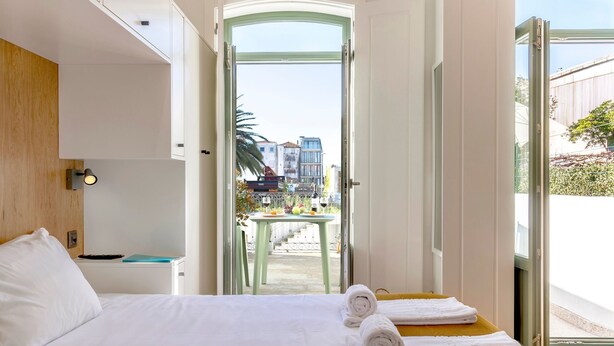 A hotel room with a bed, a desk, a chair, and a view of the city through a glass door. - WALAA Home@Almada Terrace by LovelyStay (Porto)