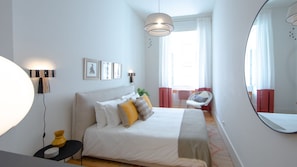 A bedroom with a bed, a chair, a desk, a mirror, and a window with curtains.