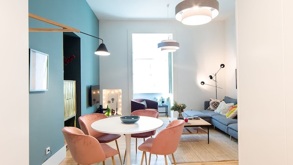 A modern living room with a round table and chairs, a sofa, and a coffee table. There is a large window providing natural light.
