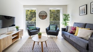 A modern living room with a grey sofa, two armchairs, a round coffee table, a TV, and a large window with a view of trees.