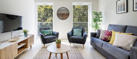 Una sala de estar moderna con un sofá gris, dos sillones, una mesa de centro redonda, un televisor y un ventanal con vistas a árboles.