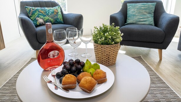 A dining table with a bottle of Mateus wine, glasses, a plate of muffins, grapes, and a small plant.