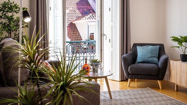 Un salon moderne avec un canapé, un fauteuil, une petite table sur laquelle se trouvent une bouteille et des verres, et une vue sur un balcon doté d’un toit en tuiles.