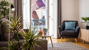 A modern living room with a sofa, a chair, a small table with a bottle and glasses, and a view of a balcony with a tiled roof.