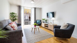 Un salon moderne avec deux canapés gris, une table basse, un téléviseur et un balcon donnant sur un immeuble jaune.