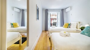 Ein Hotelzimmer mit zwei Betten, einem kleinen Nachttisch mit Lampe und Blick auf ein Gebäude durch ein Fenster.