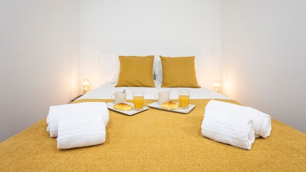 A neatly made bed with a yellow bedspread, two pillows, and two towels folded at the foot of the bed. There are two plates with croissants and two glasses of orange juice on the bed.