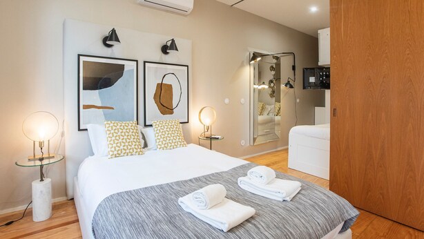A modern hotel room with a large bed, two bedside tables with lamps, a mirror, a shower area, and a TV. - Cute & Bright Flat w/ AC by LovelyStay (Porto)