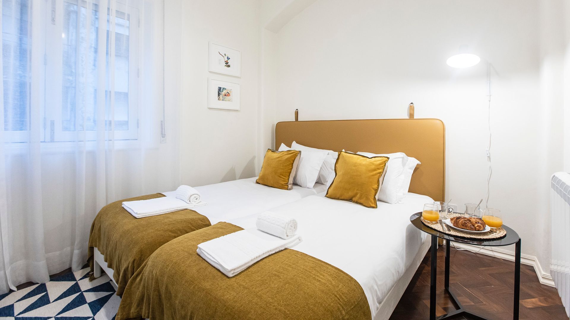 Une chambre d’hôtel comprenant un lit, deux tables de chevet, une fenêtre avec des rideaux légers et une petite table avec des articles pour le petit-déjeuner.