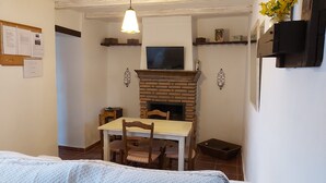 A cozy room with a fireplace, a dining table with chairs, a television above the fireplace, and a bed with a white bedspread. - Cottage (Tózar)