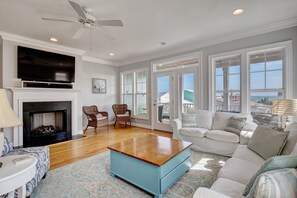 A spacious living room with a fireplace, a flat-screen TV, wicker chairs, a white sofa, and large windows offering a view of the sea.