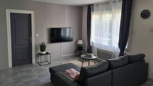 En moderne stue med en grå sofa, et fladskærms-tv, et lille sofabord med en lampe og et vindue med gardiner.