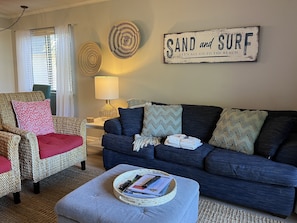 A living room with a blue sofa, wicker armchair, and ottoman. There's a sign on the wall that reads "SAND and SURF". - Comfortable, clean & well equipped 2bd 2bath - overlooking lagoon (Hilton Head Island)