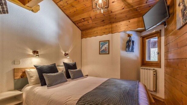 A bedroom with a wooden ceiling, a bed with pillows, a wall-mounted TV, and a window. - Verny · Appart 2CH Familiale | Pied Pistes | Piscine | Vue (Les Houches)