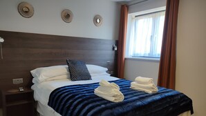 Ein ordentlich gemachtes Bett mit weißen Leinen, einer blauen Streifen-Decke und zwei Handtüchern. Es gibt ein hölzernes Kopfende, einen Nachttisch mit einer Lampe und ein Fenster mit Vorhängen.