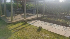 Ein überdachter Außenbereich mit Stühlen und Tischen, umgeben von einem Garten und Bäumen.