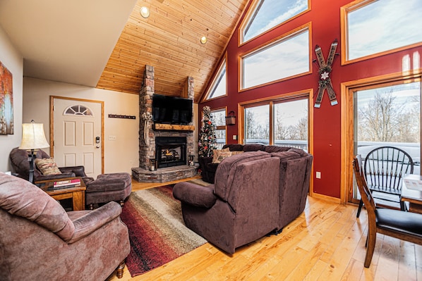 A cozy living room with a fireplace, wooden floors, and large windows.