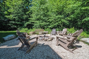 An outdoor seating area with Adirondack chairs and a fire pit surrounded by rocks and gravel, set against a backdrop of dense green trees.