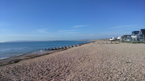 A pebbled beach with a wooden breakwater, houses along the shore, and a clear blue sky. - Charming 3-bed Caravan in Camber Food Rest (Rye)