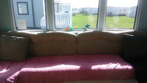 A room with a beige sofa against a window overlooking a grassy yard. - Charming 3-bed Caravan in Camber Food Rest (Rye)
