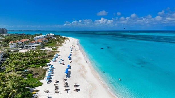 Ein Strand mit klarem türkisblauem Wasser, einer Reihe von Sonnenschirmen und Gebäuden im Hintergrund.