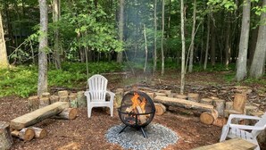 A forested area with a fire pit, white chairs, and logs arranged around it. - S'more Fun by Taylor Made Deep Creek (Oakland)