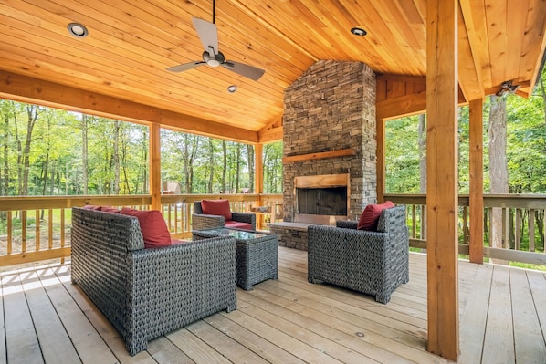 A covered outdoor deck with wicker furniture, a stone fireplace, and a wooden ceiling.