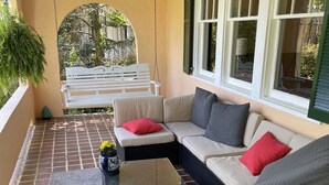 Une terrasse ensoleillée, meublée de mobilier en osier, avec une balançoire et une vue sur l’extérieur.