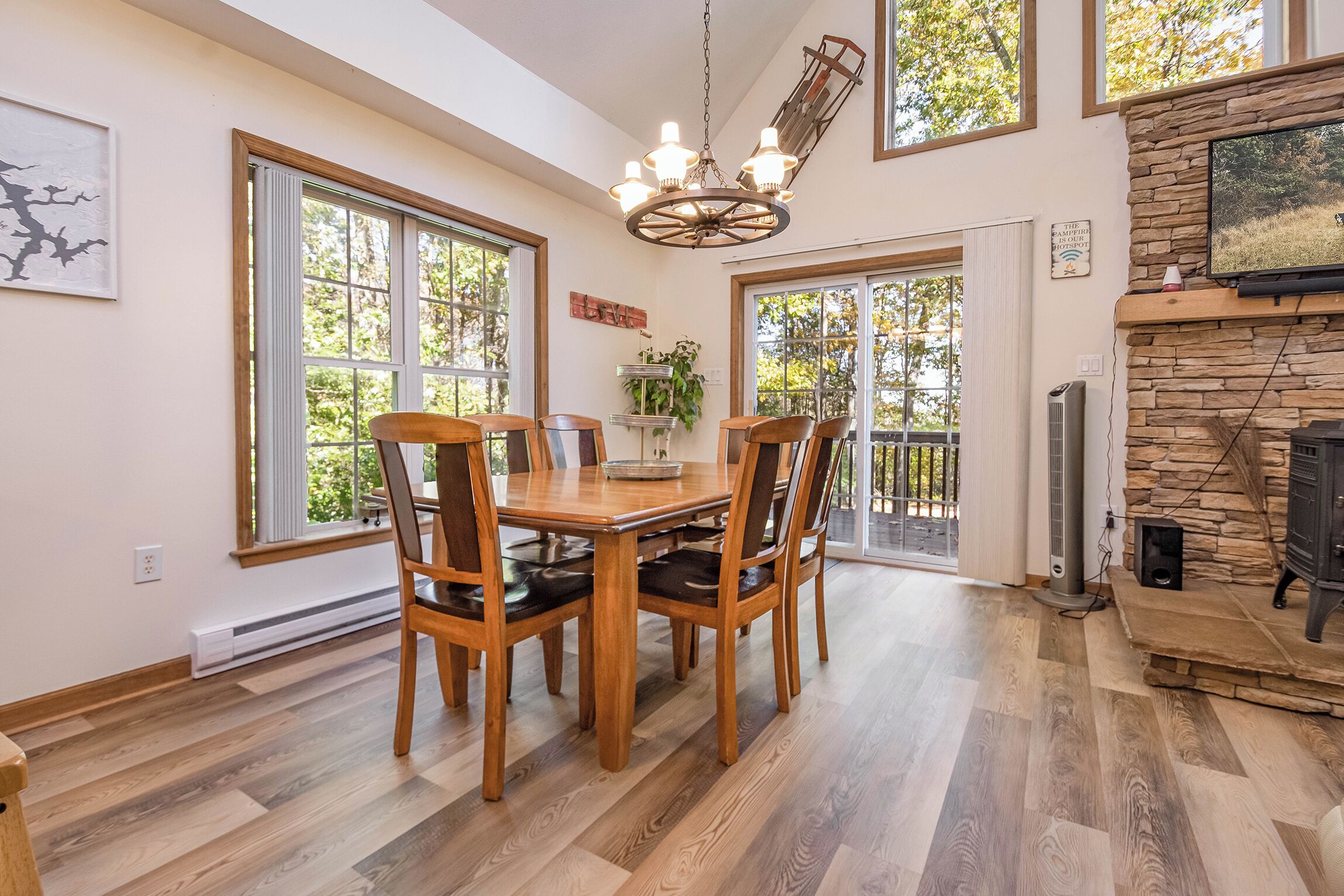 A dining area with a wooden table and chairs, large windows, a stone fireplace, and a mounted television.