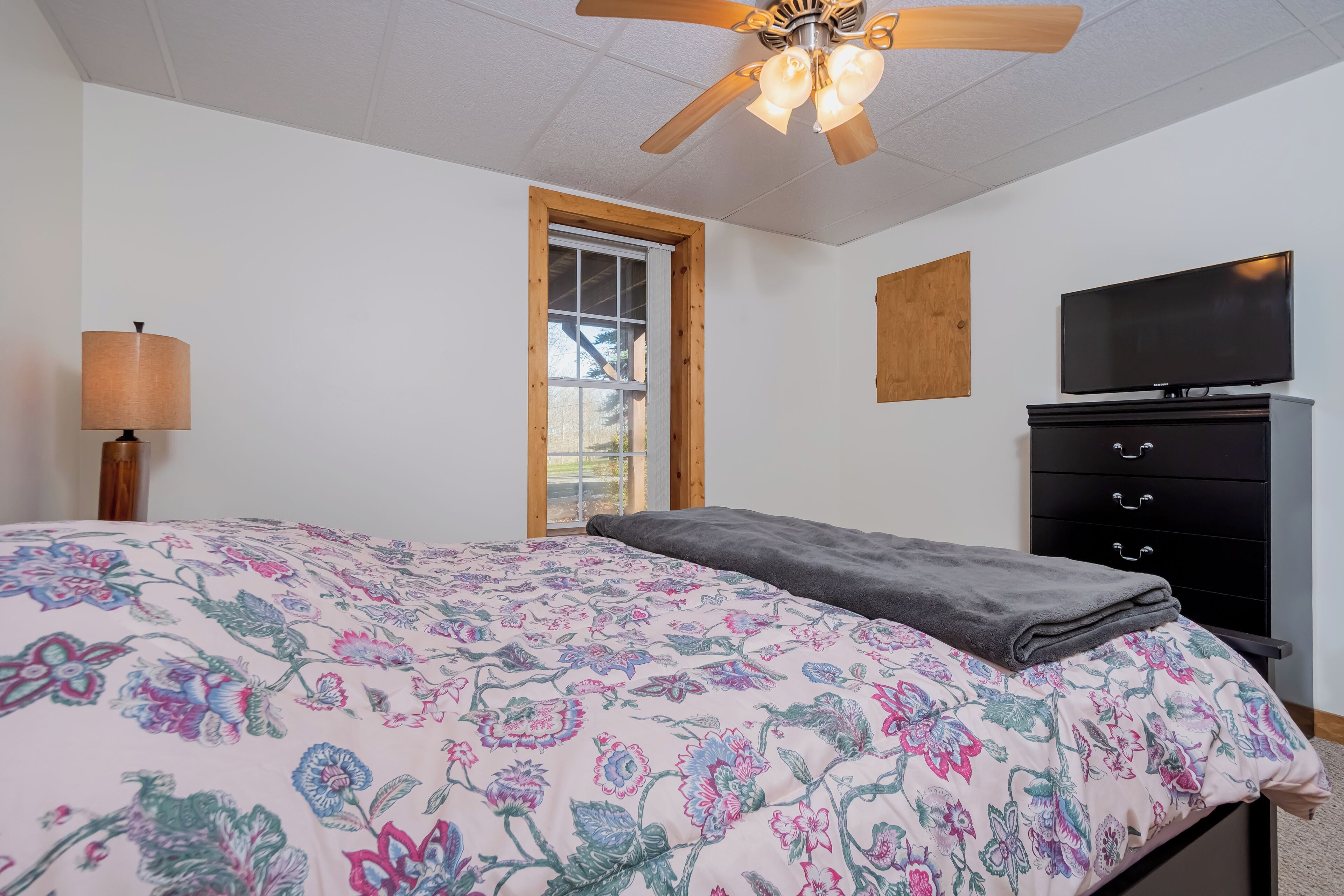 A bedroom with a bed, a ceiling fan, a TV, a dresser, and a window.