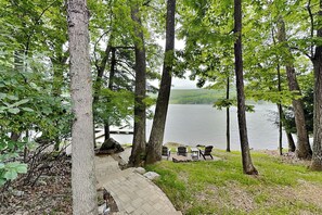 A lakeside view with a paved walkway, wooden chairs, and a stone bench.