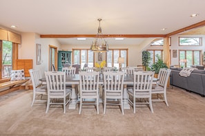 A spacious dining area with a large wooden table surrounded by white chairs, a chandelier, and large windows providing natural light.