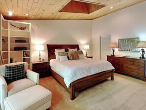 Ein Schlafzimmer mit einem hölzernen Bett, weißer Bettwäsche, einem weißen Sessel, einer Holzkommode und einem Bücherregal.