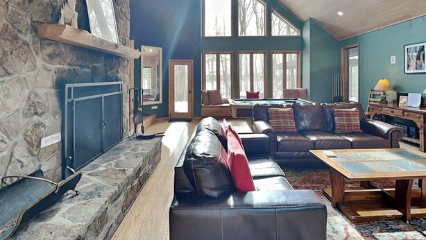 A spacious living room with a stone fireplace, leather sofas, a pool table, and large windows allowing natural light.