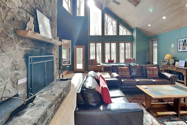 A spacious living room with a stone fireplace, leather sofas, a pool table, and large windows allowing natural light.